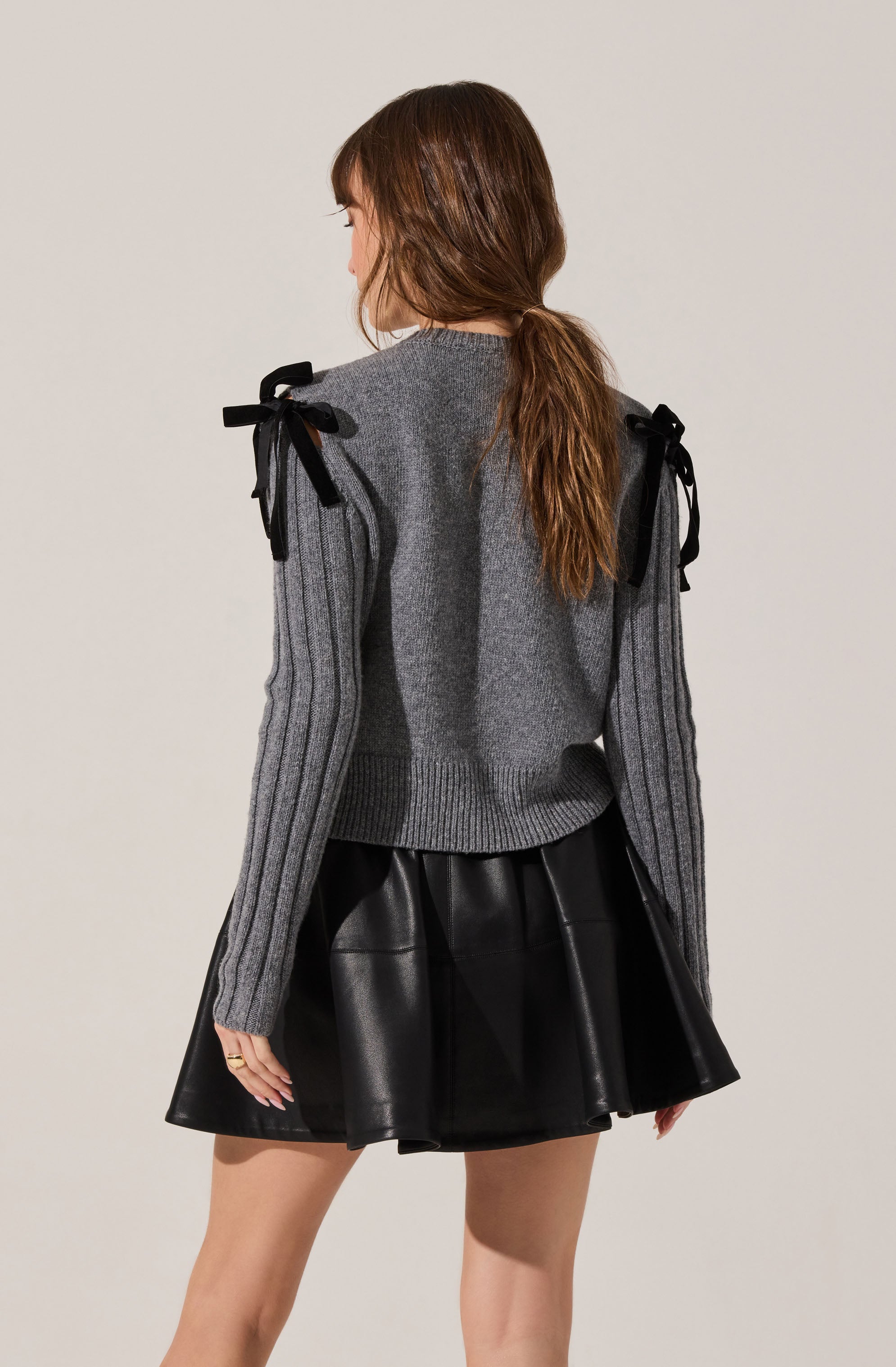 A woman is wearing a gray knit sweater with ribbed details on the sleeves and shoulder bows made of black ribbon. She pairs the sweater with a black, shiny, leather-like skirt.