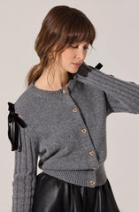 A woman is wearing a grey knit cardigan with gold heart-shaped buttons down the front. The cardigan has ribbed cuffs and hem, and features black ribbon ties on the shoulders. She is also wearing a black skirt made of a shiny, leather-like material.