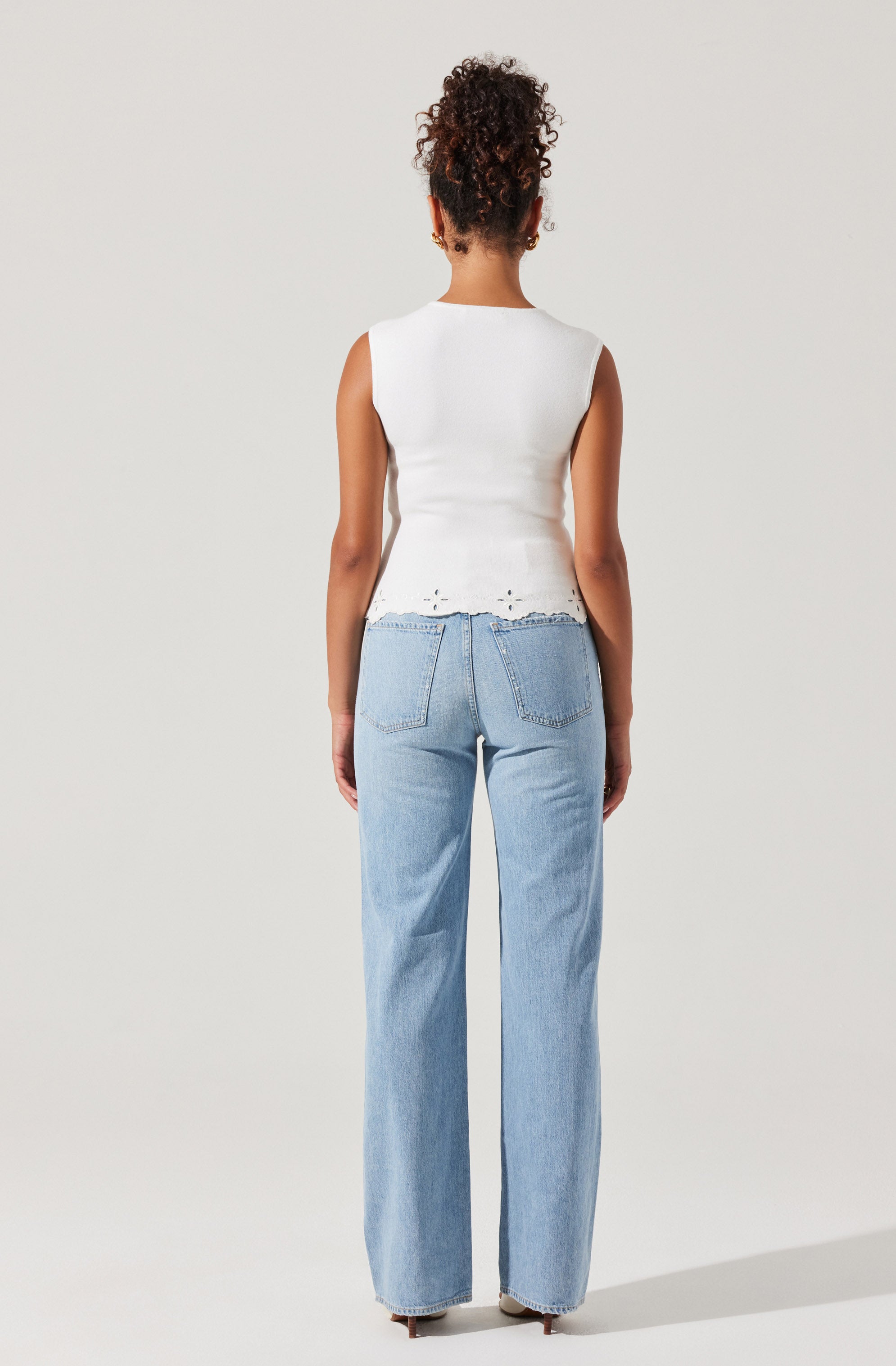 A woman is wearing a sleeveless white top with frayed edges at the hem, paired with light blue high-waisted, wide-leg jeans.