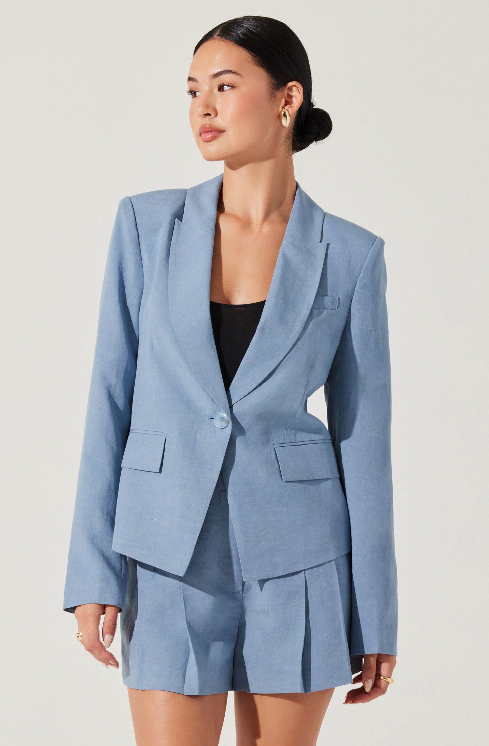 A woman is wearing a light blue tailored blazer with peaked lapels, a single button closure, and flap pockets. She pairs it with matching high-waisted shorts featuring pleats. She has dark hair styled in a low bun and is accessorized with gold hoop earrings and rings.