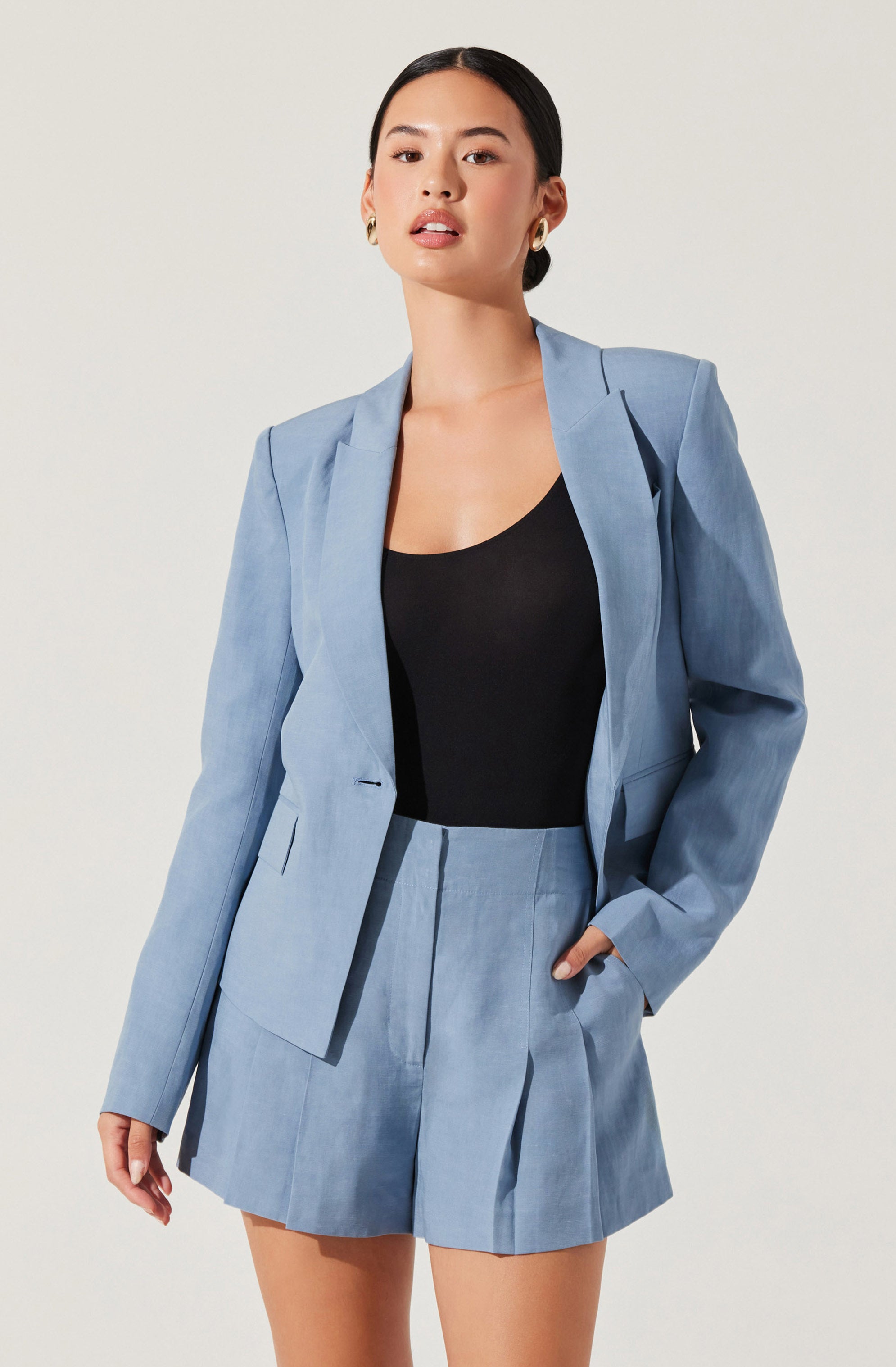 A woman is wearing a light blue tailored suit consisting of a blazer and matching high-waisted shorts. The blazer has structured shoulders, a notched lapel collar, and is slightly fitted. The shorts have a pleated front and a waistband, with one hand casually tucked into the pocket. She also wears a black sleeveless top underneath the blazer.