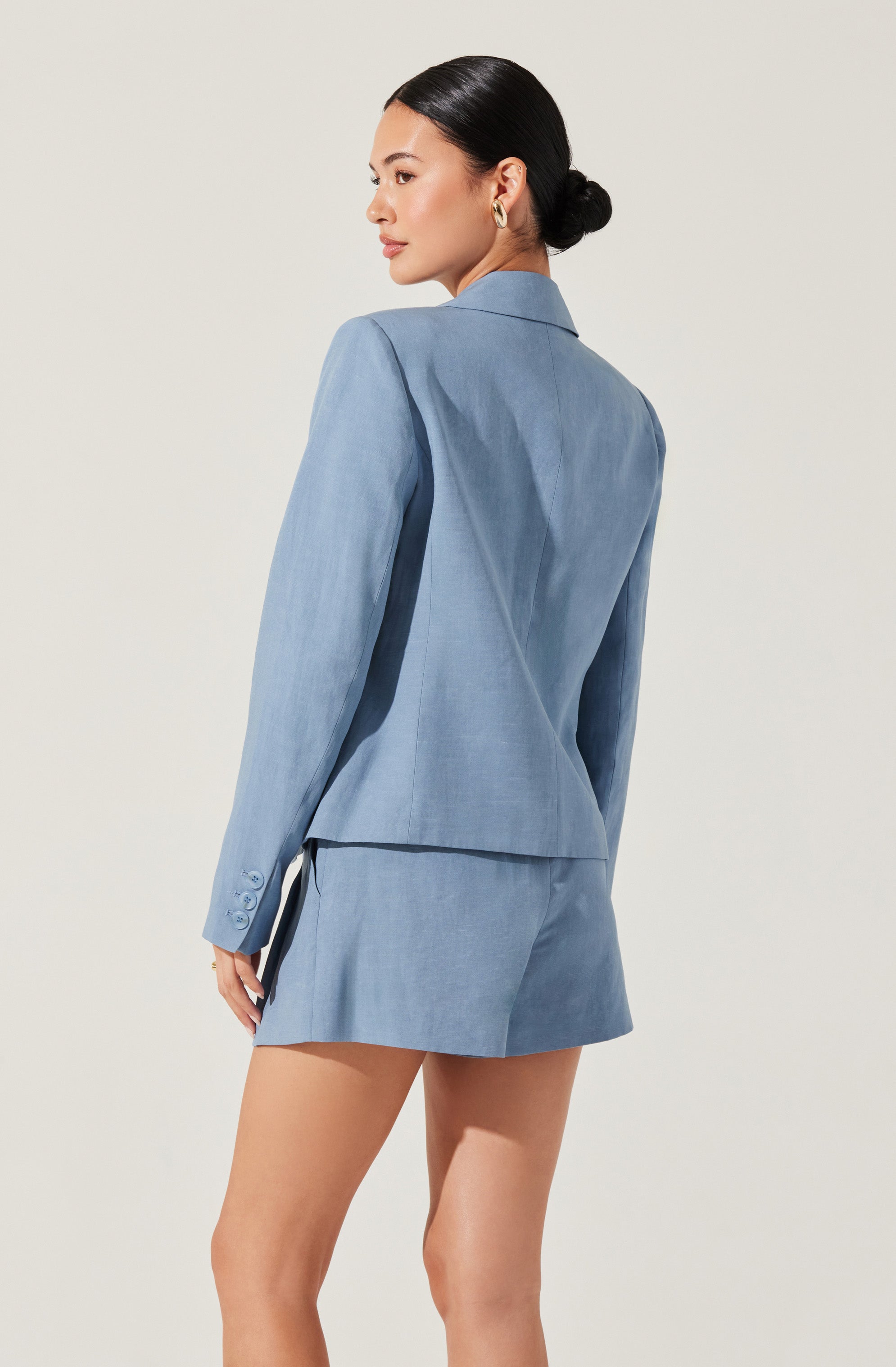 A woman is wearing a light blue blazer with long sleeves and a notched lapel collar. The blazer is tailored, fitted at the waist, and features three buttons on each sleeve cuff. She pairs it with a matching light blue skirt that has a subtle slit on the side.
