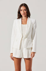 A woman is wearing a white tailored blazer with a single-button closure, structured shoulders, and flap pockets. She pairs it with matching high-waisted white shorts that have a loose fit and pleats. Underneath, she has on a cream-colored, textured sleeveless top.