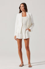 A woman is dressed in an all-white outfit consisting of a tailored blazer with long sleeves and notched lapels, a sleeveless textured top, and high-waisted shorts with pleats. She is also wearing strappy white sandals.