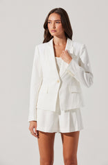 A woman is wearing a white, tailored blazer with a single button closure and notched lapels. Underneath, she has on a light-colored, possibly knitted top. She is also wearing matching white shorts with a high waist and pleats, creating a polished yet casual look.