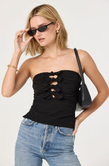 A woman wearing a strapless black top with ruched fabric and bow details down the front. She pairs it with light blue high-waisted jeans. She accessorizes with small gold earrings, a gold bracelet, and black sunglasses. She carries a black shoulder bag.
