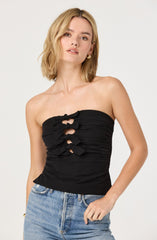 A woman is wearing a strapless black top with ruched fabric and decorative cutouts in the front, featuring tied bow details. She is also wearing light-wash denim jeans.