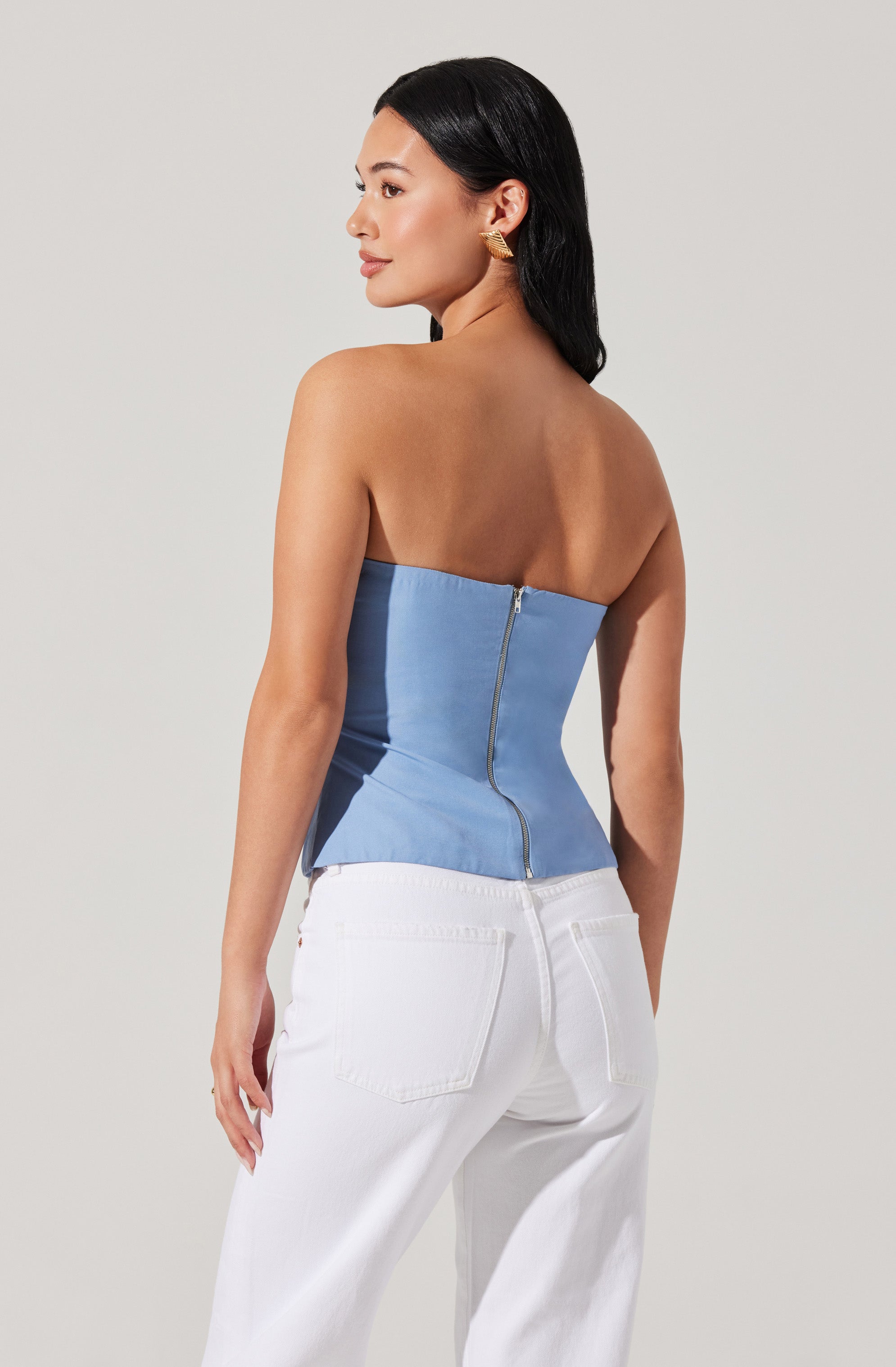 A woman is wearing a strapless, light blue corset top with a visible zipper running down the back, paired with high-waisted white jeans that have back pockets.