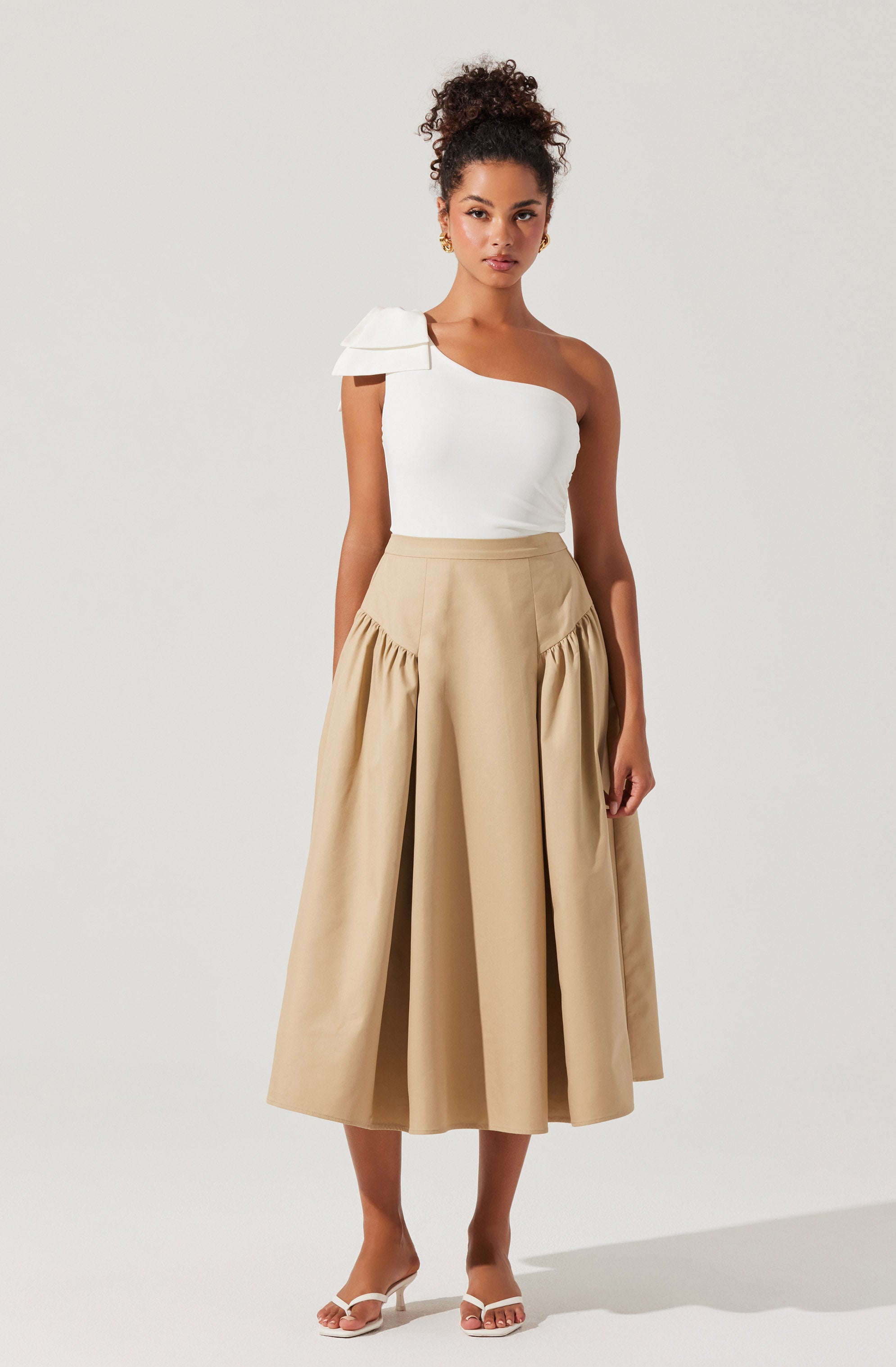 A woman is wearing a one-shoulder white top with a layered ruffle detail on the shoulder, paired with a high-waisted beige midi skirt featuring gathered fabric at the hips and wide pleats. She is also wearing open-toe sandals with a block heel.