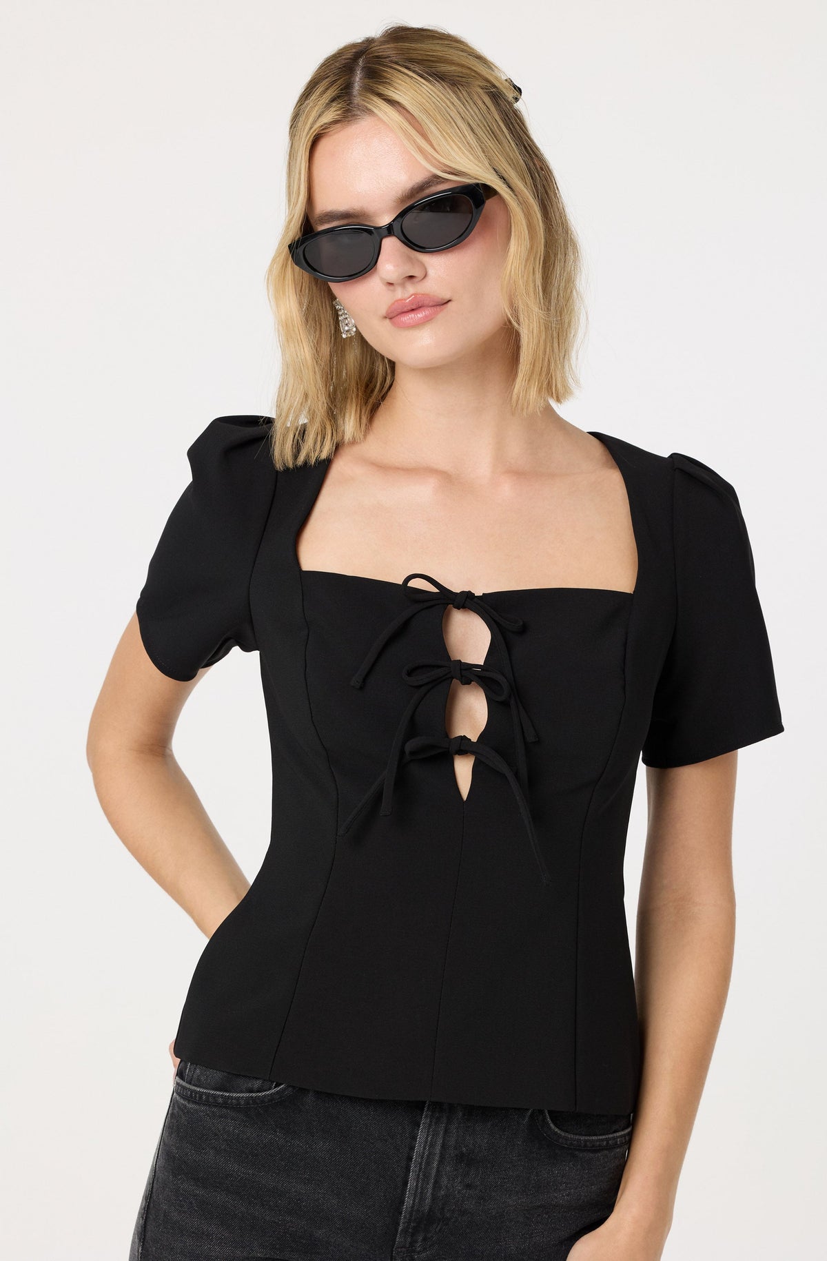 A woman is wearing a black, fitted short-sleeve top with puffed sleeves and a square neckline. The top features a distinctive front detail with three tied bows in the center. She has blonde, wavy hair and is accessorizing with large, black sunglasses and drop earrings.