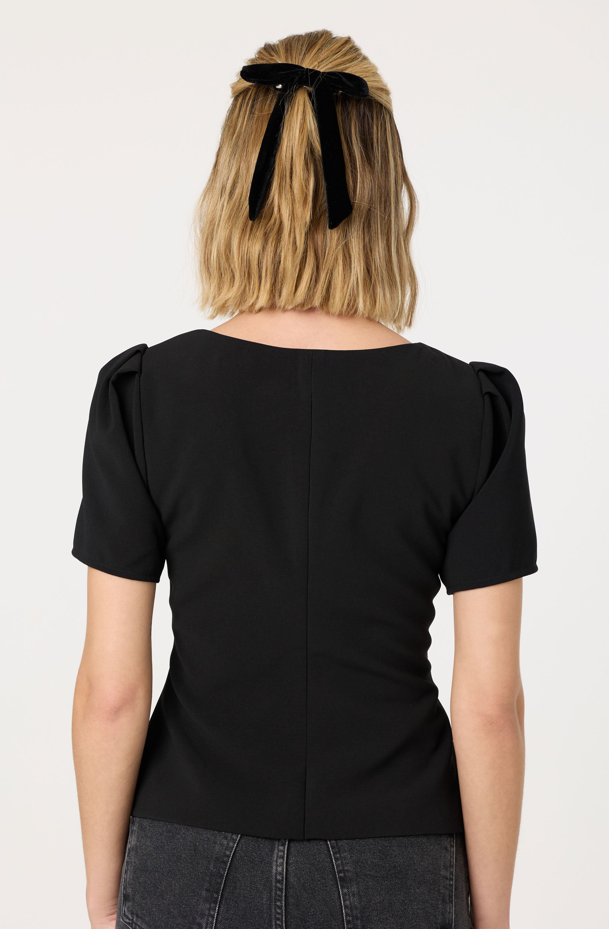 A woman is wearing a black short-sleeved top with puffed shoulders and a round neckline. She has a black ribbon tied into a bow in her blonde hair.