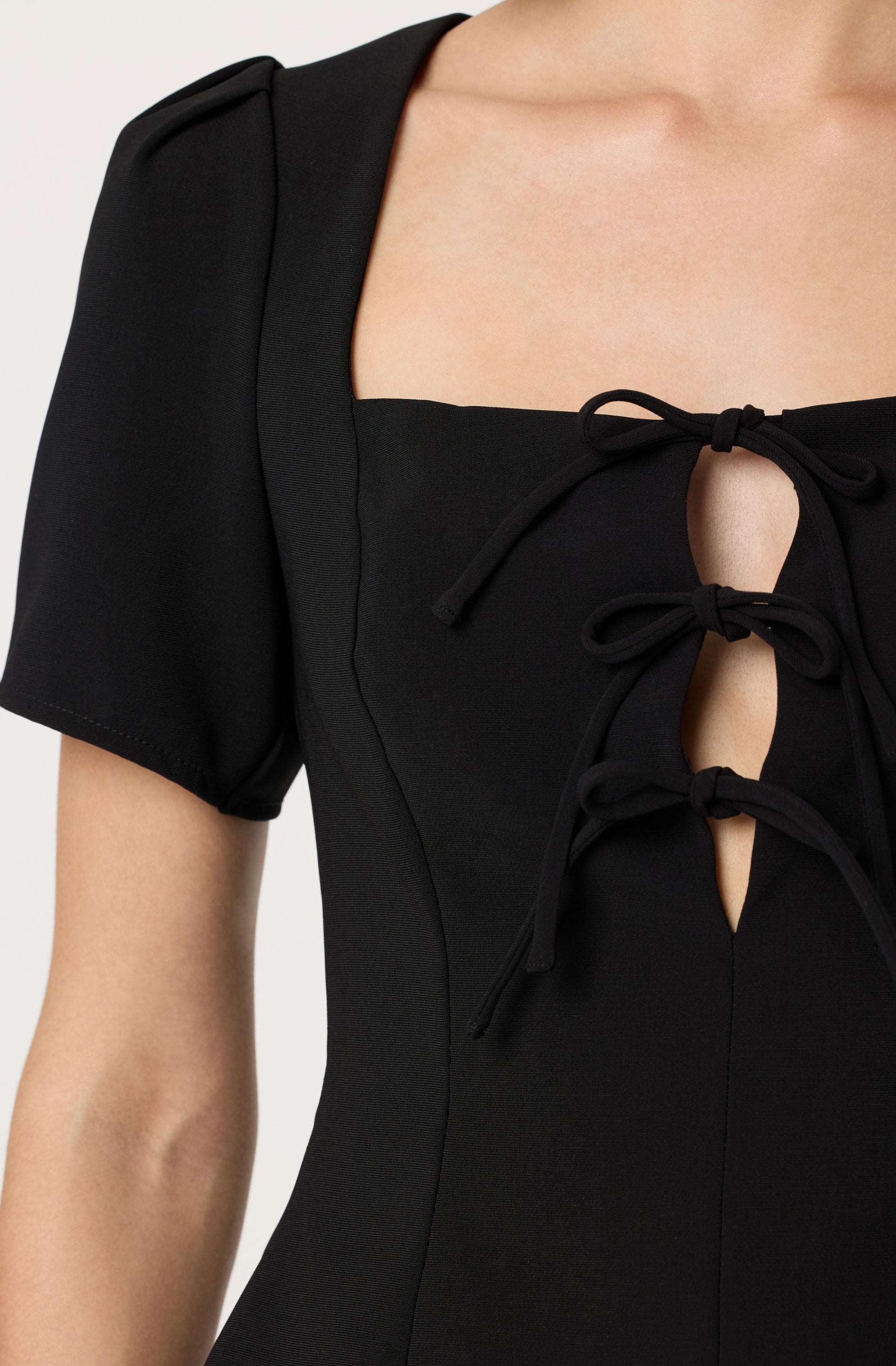 A close-up of a black dress featuring short puff sleeves and a square neckline with three decorative ties that create keyhole openings down the front.