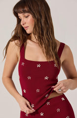 A woman is wearing a sleeveless, deep red top with wide straps. The top features small white floral patterns evenly distributed across the fabric. She is also wearing a matching deep red skirt with the same white floral patterns.