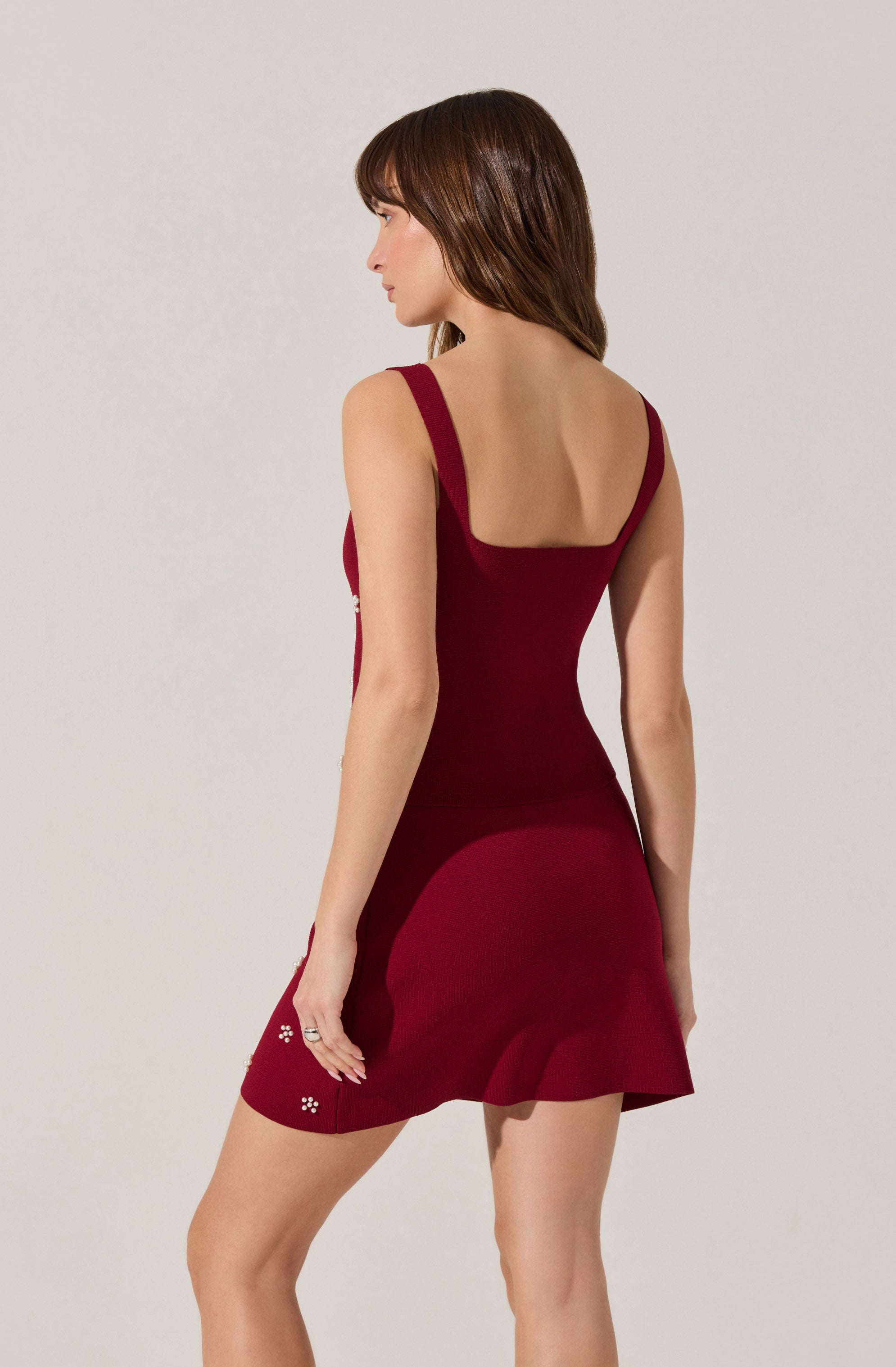 A woman is wearing a sleeveless, fitted, deep red dress with wide shoulder straps and a square neckline. The dress features a ruffled hemline and decorative white flower embellishments near the bottom.