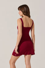 A woman is wearing a sleeveless, fitted, deep red dress with wide shoulder straps and a square neckline. The dress features a ruffled hemline and decorative white flower embellishments near the bottom.