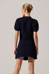 A woman is wearing a matching set consisting of a navy blue textured top with puffed short sleeves and a matching skirt. The top has a ribbed, wave-like pattern, and the skirt features the same textured pattern with a slightly flared hem. She has her hair styled in a sleek, low bun and is wearing earrings.