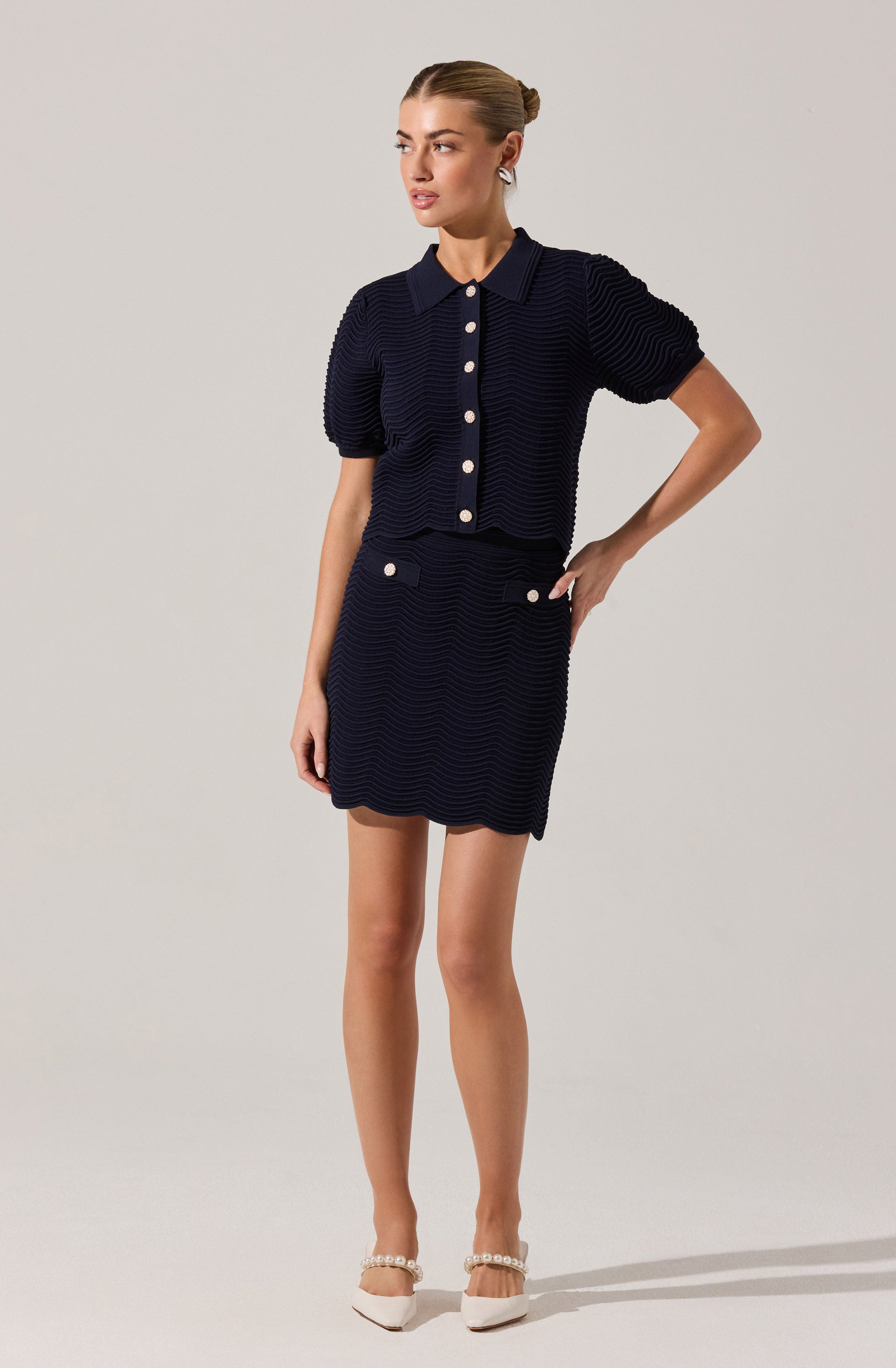 A woman is wearing a matching navy blue textured outfit consisting of a short-sleeved, collared button-up top and a high-waisted skirt. The top has large white buttons down the front, and the skirt features two white buttons near the pockets. She is also wearing white shoes with pearl embellishments on the strap.