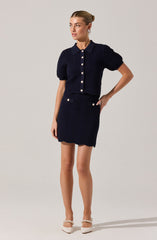 A woman is wearing a matching navy blue textured outfit consisting of a short-sleeved, collared button-up top and a high-waisted skirt. The top has large white buttons down the front, and the skirt features two white buttons near the pockets. She is also wearing white shoes with pearl embellishments on the strap.