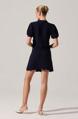The woman is wearing a matching navy blue textured two-piece outfit consisting of a top with short, puffed sleeves and a high neckline, and a matching skirt with a slightly flared hem. She is also wearing light-colored high-heeled shoes.