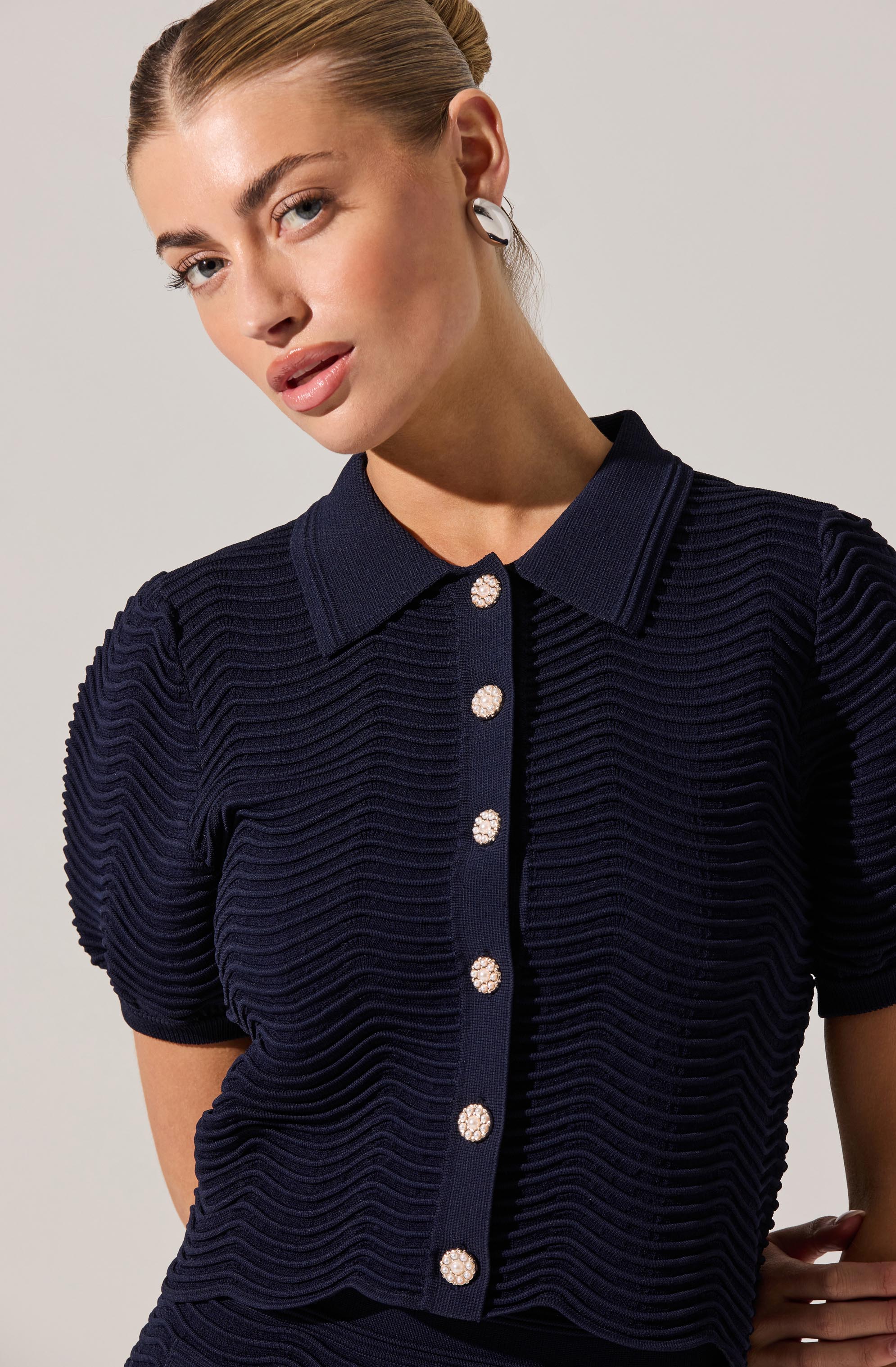 A woman is wearing a navy blue textured short-sleeve button-up top with a collar. The top features a ribbed, wavy pattern across the fabric, with decorative shiny, pearl-like buttons down the front.