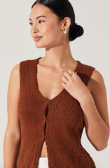 A woman wearing a sleeveless, rust-colored, textured knit top with a deep V neckline and a front opening. She has dark hair styled in a low bun, and she is accessorized with gold hoop earrings and a gold ring on her right hand.