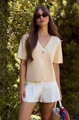 Woman wearing a cream-colored, short-sleeved V-neck top with a single large button at the front, paired with white shorts. She has long, wavy brown hair, is wearing dark sunglasses, and is standing outdoors amid lush green foliage.