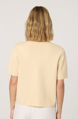 A woman is wearing a short-sleeved, beige top made of a textured, knit-like fabric. The top has a simple, round neckline and a loose fit. She is also wearing a light-colored skirt.
