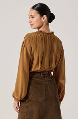 A woman is wearing a long-sleeved, silky, brown blouse with a gathered neckline and pleated details on the back. The blouse has slightly puffed sleeves with elastic cuffs. She is also wearing high-waisted, brown corduroy pants with front and back pockets.