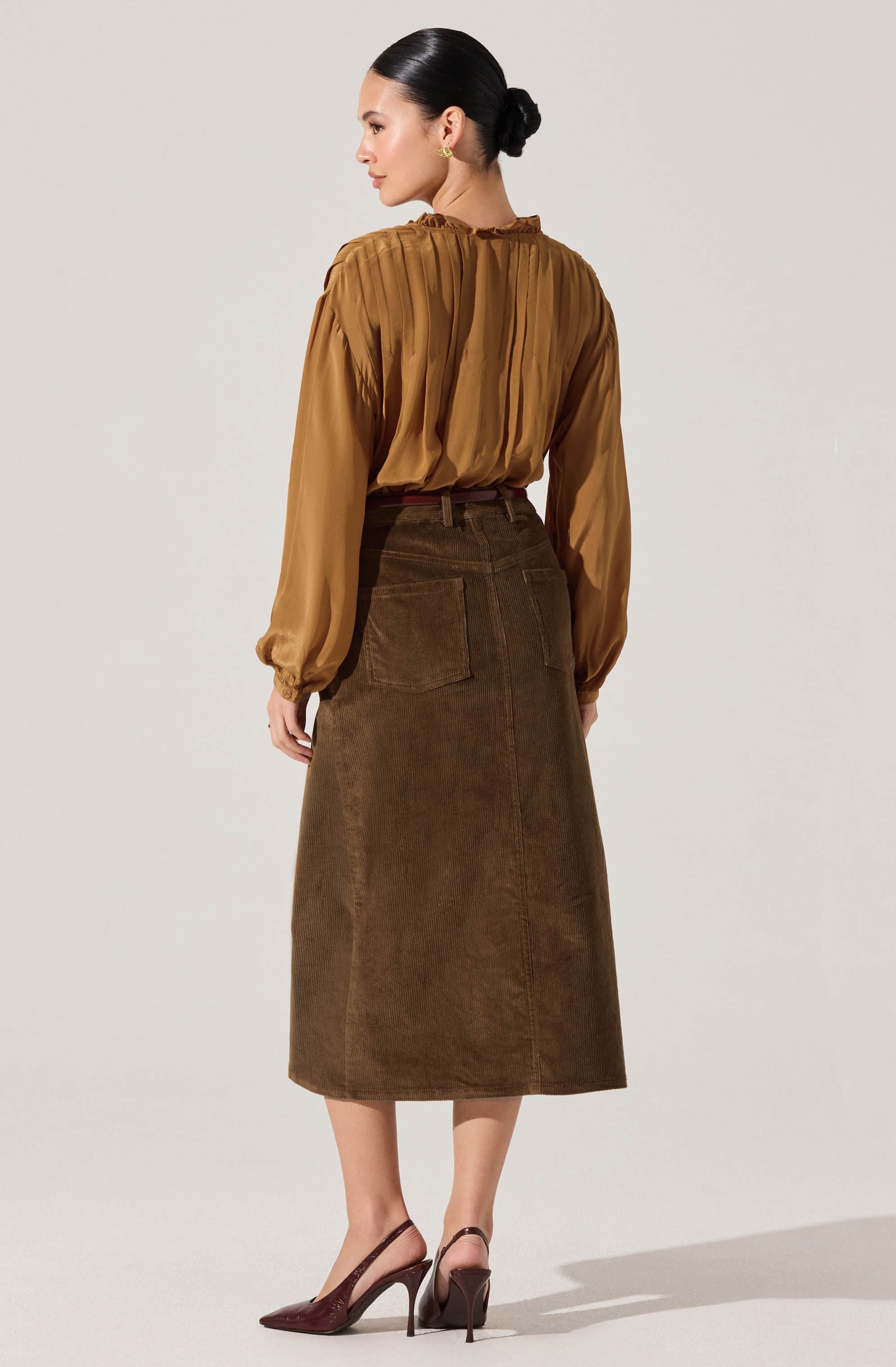 A woman is wearing a long-sleeved, mustard-colored blouse with a pleated design and a high neckline. She pairs it with a high-waisted, dark brown corduroy A-line skirt that reaches mid-calf, featuring large back pockets. She completes the outfit with burgundy high heel shoes.