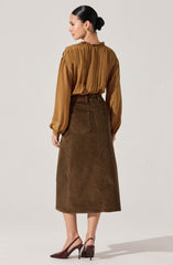 A woman is wearing a long-sleeved, mustard-colored blouse with a pleated design and a high neckline. She pairs it with a high-waisted, dark brown corduroy A-line skirt that reaches mid-calf, featuring large back pockets. She completes the outfit with burgundy high heel shoes.