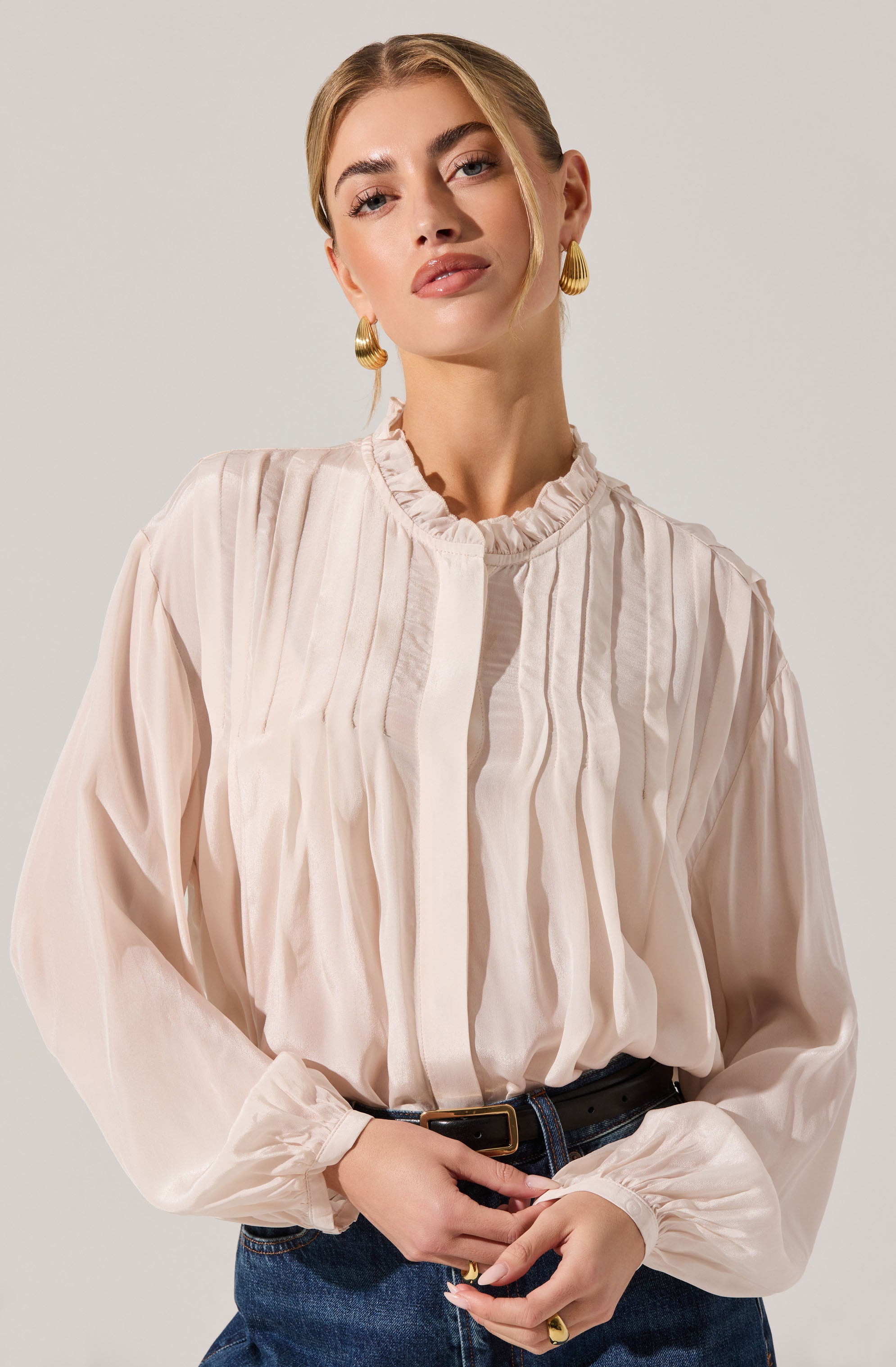 A woman is wearing a light-colored, long-sleeved blouse made of a sheer, flowy fabric. The blouse features vertical pleats on the front, a high ruffled neckline, and gathered cuffs at the wrists. She has paired the blouse with dark jeans and a black belt. She accessorizes with large gold hoop earrings and a gold ring, her blonde hair styled neatly.