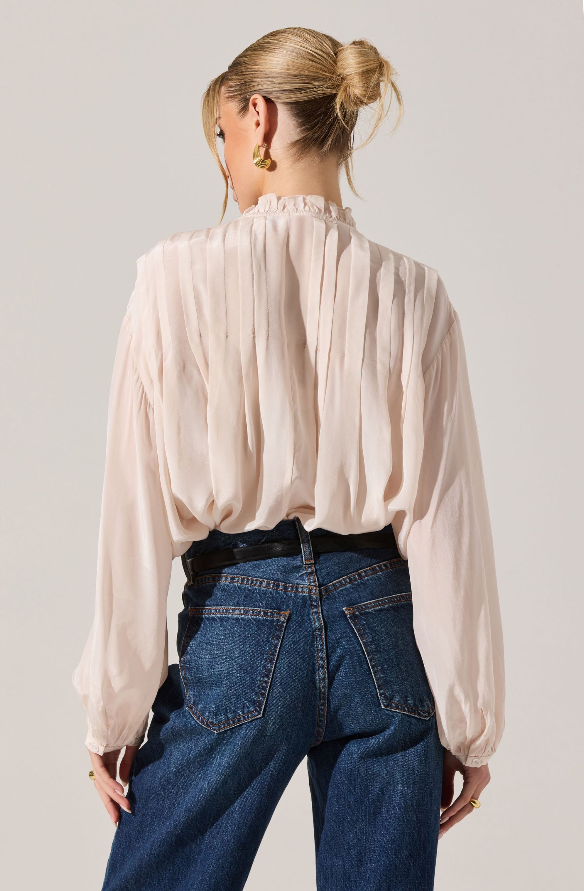A woman is wearing a light-colored, long-sleeved blouse with a high, ruffled collar and vertical pleats. The blouse is loose-fitting with gathered cuffs at the wrists. She pairs it with high-waisted, dark blue denim jeans featuring prominent back pockets. Her hair is styled in a casual, low bun, and she is accessorized with large, gold earrings.