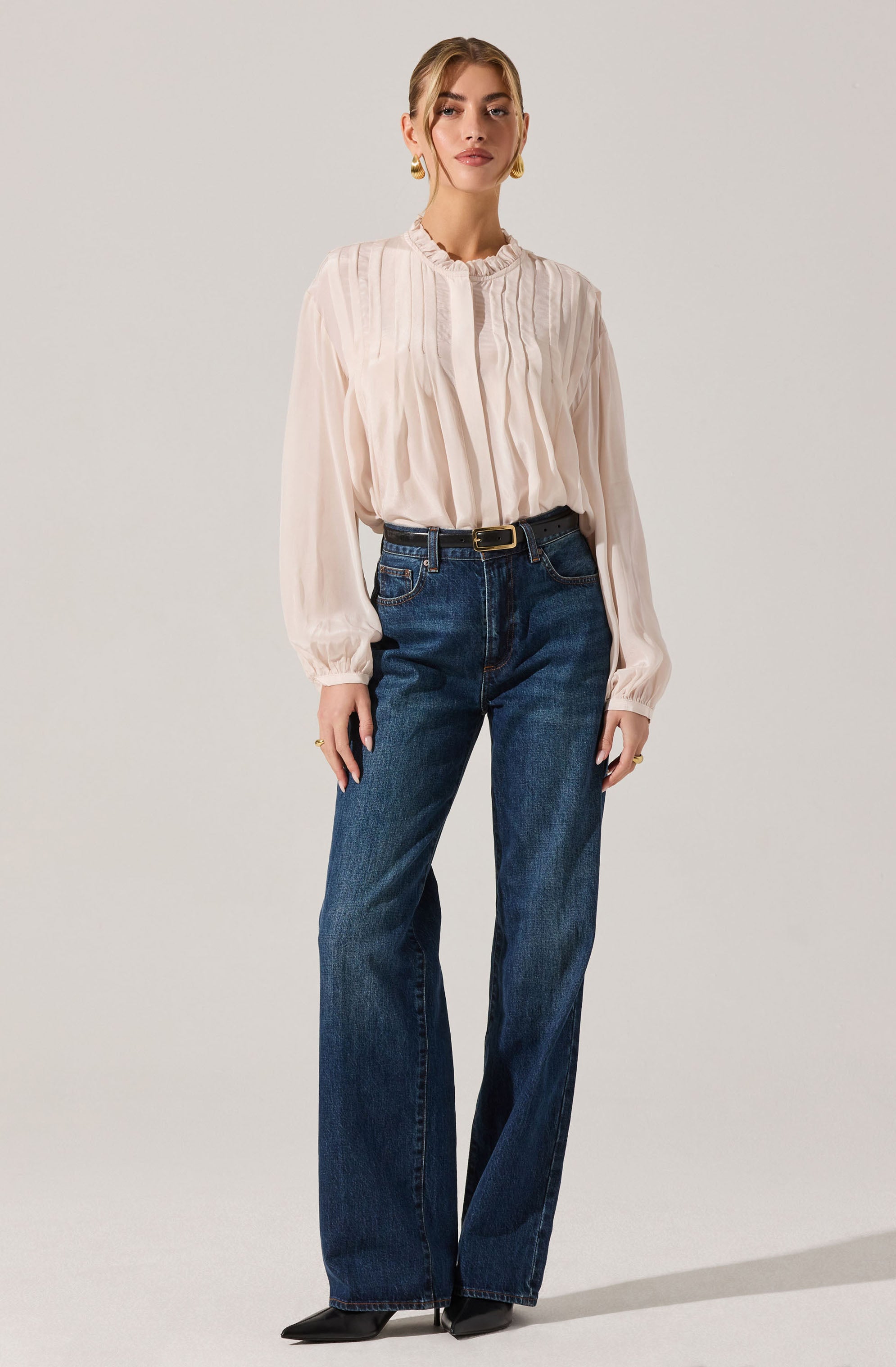 A woman is wearing a long-sleeved, light-colored blouse with vertical pleats and a ruffled neckline. She pairs it with high-waisted, dark blue wide-leg jeans that have a black belt with a gold buckle. She completes the outfit with black pointed shoes.