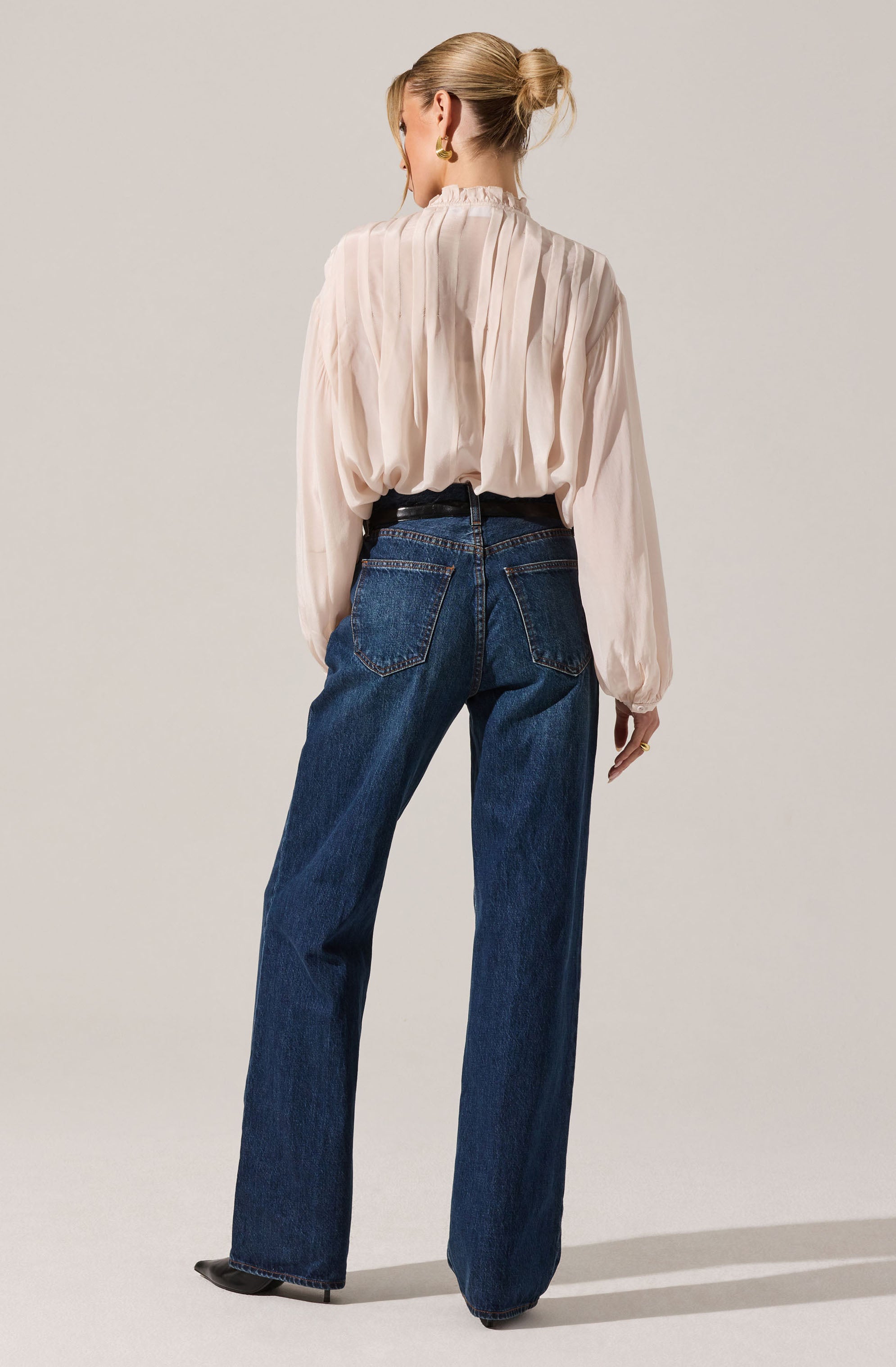 A woman is wearing a long-sleeved, light-colored, pleated blouse with a high neckline. The blouse has a slightly loose fit and is tucked into high-waisted, dark blue jeans with a straight-leg cut. The jeans feature visible stitching and back pockets. She is also wearing black shoes with a pointed toe and gold hoop earrings. Her hair is styled in a low bun.