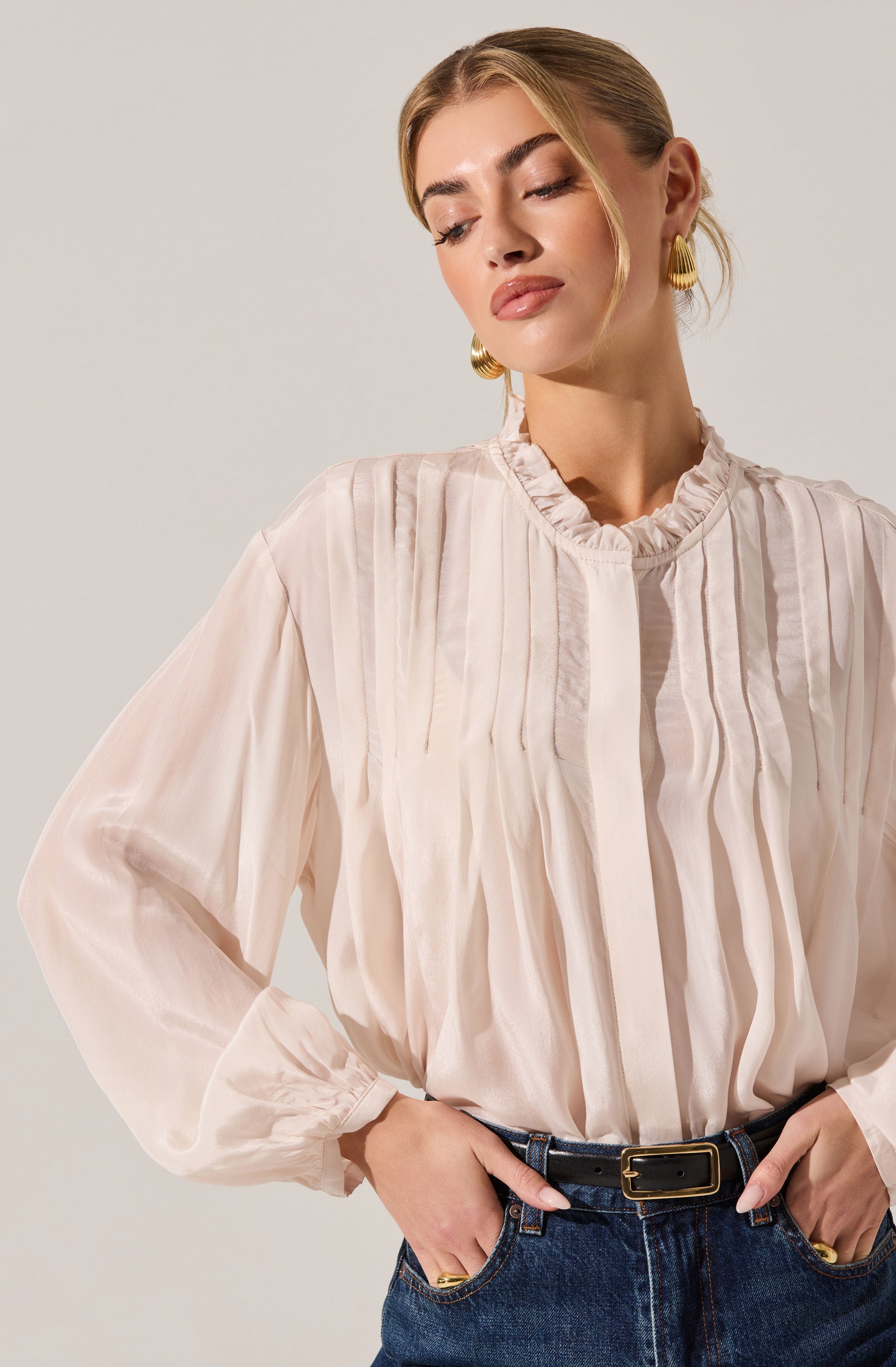 A woman is wearing a light-colored, long-sleeved blouse with vertical pleats and ruffled detailing around the collar. She has paired it with dark blue jeans and a black belt with a gold buckle. She is also wearing gold hoop earrings.