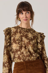 A woman is wearing a brown long-sleeve blouse with a floral print pattern in shades of beige and cream. The blouse features ruffled details along the shoulders and a gathered neckline. She is also wearing a high-waisted, brown corduroy skirt. She has gold hoop earrings and shoulder-length brown hair with bangs.