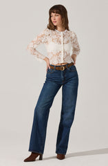 A woman is wearing a long-sleeved, cream-colored lace crop top with a floral pattern. She pairs it with high-waisted, dark blue jeans with a straight leg fit, secured with a brown belt that has a gold buckle. She completes her outfit with brown ankle boots.