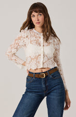 A woman is wearing a sheer, long-sleeved, button-up blouse made of white lace with floral patterns. The blouse has a high, ruffled neckline and a scalloped hem. She is also wearing blue jeans with a high waist, accessorized with a brown belt featuring a gold buckle.