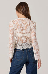 A woman is wearing a long-sleeved, sheer lace top with a floral pattern. The top is white or cream-colored, and the floral lace design is intricate, covering the entire back and sleeves. She pairs the top with high-waisted, dark blue jeans.