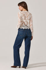 A woman is wearing a long-sleeved, white lace blouse with floral patterns, which is semi-transparent. She pairs it with high-waisted, dark blue jeans that have prominent stitching on the pockets and a relaxed fit. She is also wearing brown shoes with a pointed toe and a small heel.