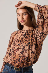 A woman is wearing a long-sleeved blouse with a round neckline, featuring a leopard print pattern in shades of brown, black, and beige. The blouse has a slightly loose fit and a gathered detail at the neckline. The fabric appears lightweight and silky. She has paired the blouse with dark blue high-waisted jeans and a black belt with a rectangular gold buckle.