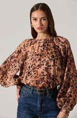 A woman is wearing a long-sleeved blouse with a leopard print pattern in shades of tan, brown, and black. The blouse has a gathered neckline with subtle pleats and voluminous sleeves that are cinched at the wrists. She is also wearing high-waisted blue jeans with a black belt.