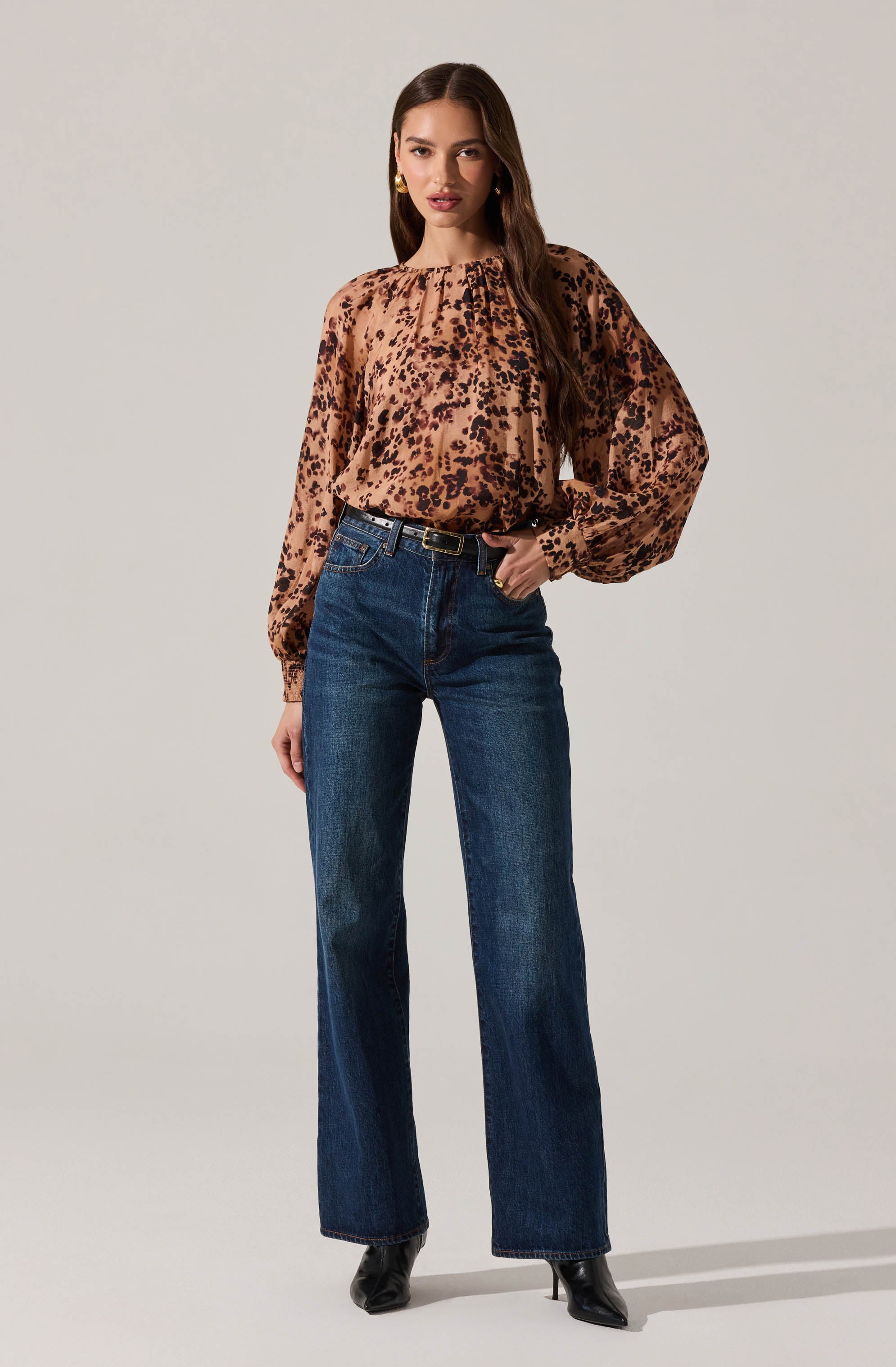 A woman is wearing a long-sleeved blouse with a leopard print pattern in shades of brown and black, and high-waisted dark blue jeans. The blouse has a loose fit with gathered detailing at the neckline and billowing sleeves. She is also wearing black pointed-toe ankle boots.