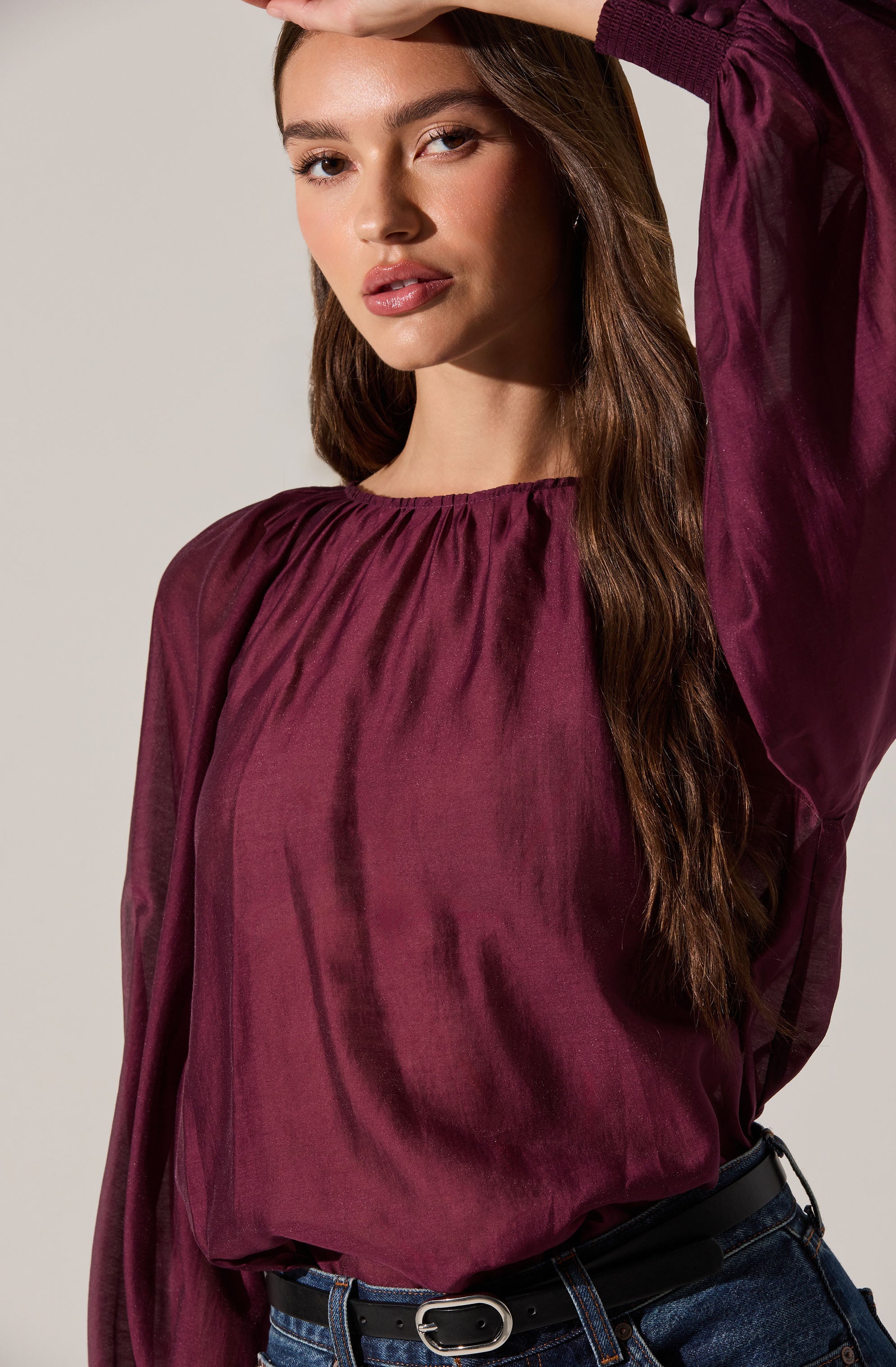 A woman is wearing a long-sleeved, shiny, deep wine-colored blouse with a gathered neckline. The blouse has a loose fit and a silky texture. She pairs it with dark blue jeans and a black belt with a silver buckle.