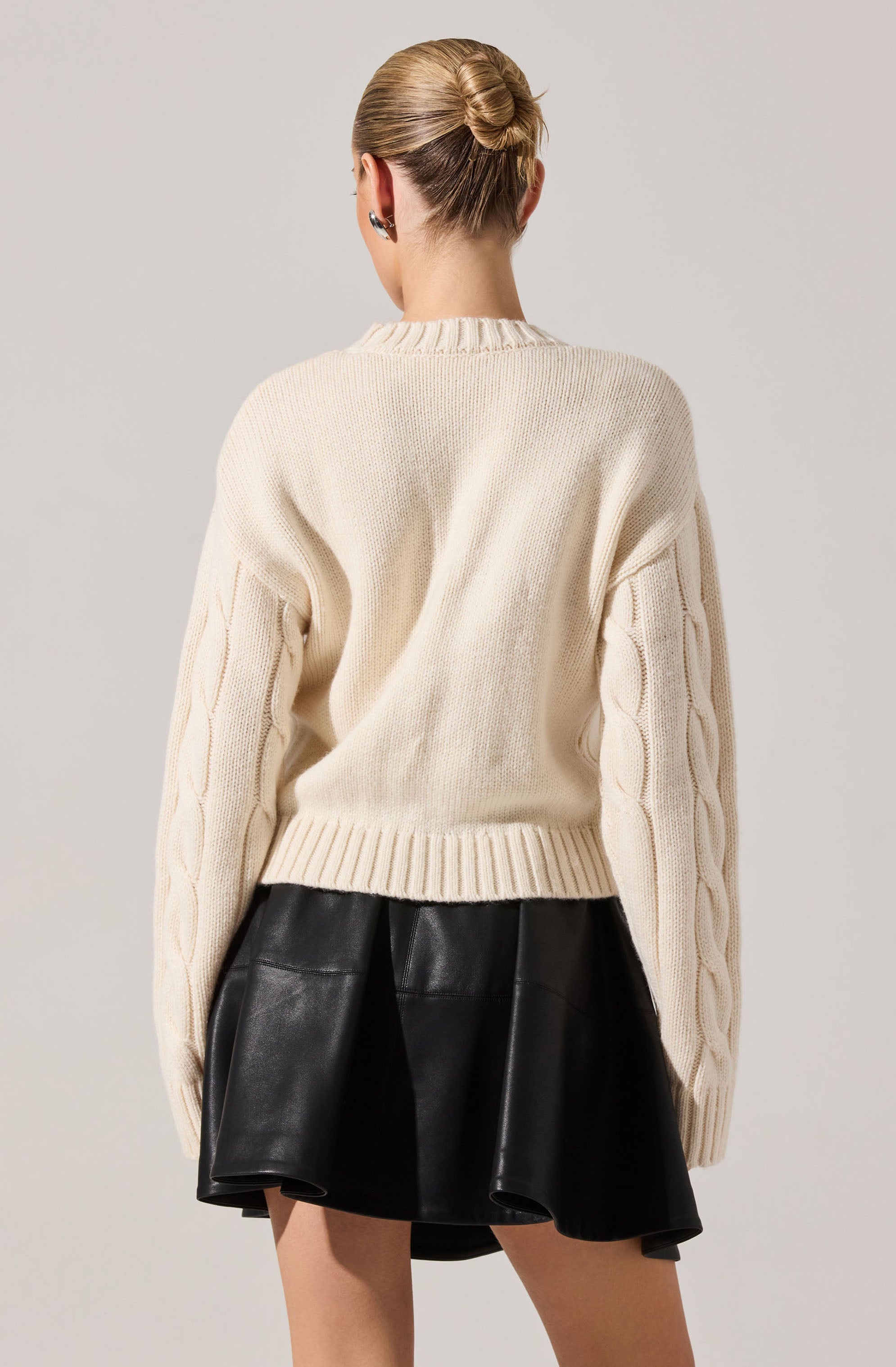 A person is wearing a chunky cream-colored knit sweater with cable knit details running down the sleeves. The sweater has a round neckline and a ribbed hem. They are also wearing a black leather skirt with a flared, A-line shape. The person's hair is styled in a neat bun.