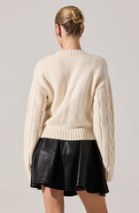 A person is wearing a chunky cream-colored knit sweater with cable knit details running down the sleeves. The sweater has a round neckline and a ribbed hem. They are also wearing a black leather skirt with a flared, A-line shape. The person's hair is styled in a neat bun.