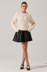 A woman is wearing an oversized, chunky knit sweater in a cream color with cable-knit details and long sleeves. She pairs it with a short, black leather flared skirt and black high-heeled shoes with ankle straps.