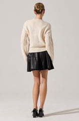 A woman is wearing an oversized, cream-colored knitted sweater with a crew neckline and cable-knit detailing along the sleeves. She pairs it with a high-waisted, black leather skirt that has a flared silhouette. She completes the outfit with black, high-heeled shoes with ankle straps.
