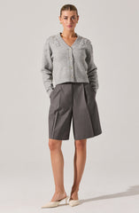 A woman is wearing a light gray, knitted cardigan with a v-neckline and floral embellishments on the shoulders. The cardigan has pearl-like buttons down the front. She is also wearing high-waisted, wide-leg gray shorts with pleats and front pockets. She has on white pointed-toe heels.
