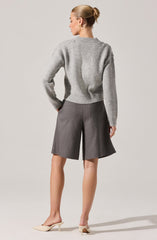 A woman is wearing a light gray, long-sleeved, slightly oversized knit sweater with a ribbed hem and cuffs. She pairs it with high-waisted, wide-leg gray shorts with a subtle texture, featuring a front crease and side pockets. She completes the look with beige pointed-toe high-heeled shoes.